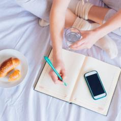 Cropped photo of crossed legs of person on bed with breakfast writing in scheduling book