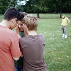 Three boys whispering in huddle with a girl in the distance