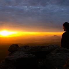 A woman sits on a cliff and stares into the sunset