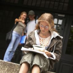 Blonde preteen sits on school steps reading while kids behind point