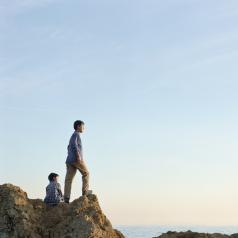 Man stands on rock cliff looking out to see while child sits behind him
