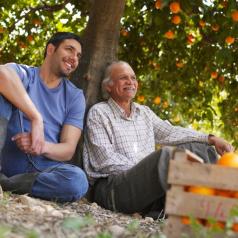 A senior and younger man sit under orange tree similing