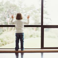 Standing child looks out of window, hands on glass