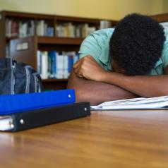Teen hides face in crossed arms on table, binders and books open nearby