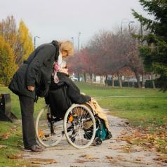 Person looks down to check on older person in wheelchair outdoors on windswept path