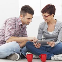 Two people sit on floor and look over schedule on tablet. A red mug sits in front of each person.