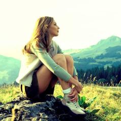 Person with long hair wearing jacket and shorts sits alone on mountaintop looking into distance