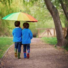 Two boys in blue jackets and rain boots walk together under rainbow umbrella