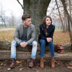 Couple sits apart on bench under tree on cold day