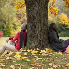 A couple sits looking away from each other on either side of a tree, with leaves scattered on the ground