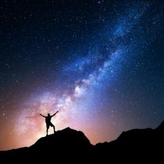 Lone backpacker stretches arms up to the night sky, in which the Milky Way is visible