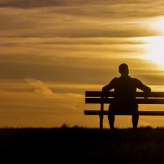 Rear view of silhouette of man sitting on bench watching the sun set in a cloudy gold sky