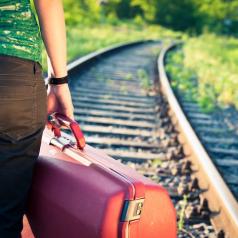 Cropped view of person holding red suitcase stands near train tracks waiting for train