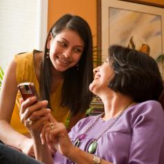 Seated, smiling mother holds up cell phone, daughter smiles and leans over to look