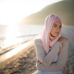 A woman who wears a hijab and has a serious expression hugs herself on the beach, looking off to the side