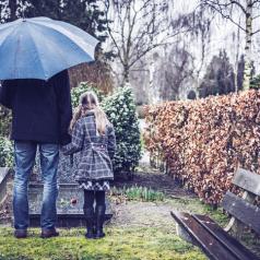 Tall adult in jeans and coat holds hand of child in dress coat with long hair as they stand at a gravestone on a rainy day