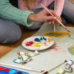 Focus on hands of mother in aqua sweater and child in pink sweater with blonde hair painting on large paper on floor