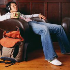 Young adult holds mug of tea and reclines in armchair wearing headphones