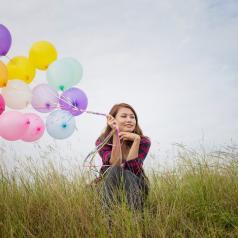 Young adult with long hair sits in tall grass, looking out into distance, holding a bunch of brightly colored balloons