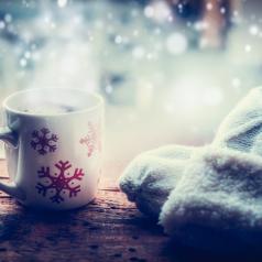 White mittents and red and white snowflake mug sit on wooden table with frosted window in background