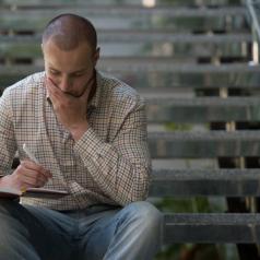 Person in button-down shirt sits on stairs, holding a pen and looking thoughtfully down into small notebook