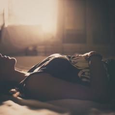 Person with short hair in tank top lies on bed, hands resting on stomach, eyes closer. Room is lit by sunset in sepia tones