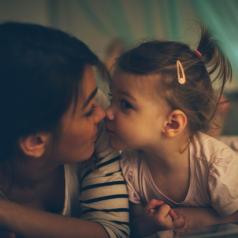 Young child and mother touch noses in dim room, gazing at each other affectionately