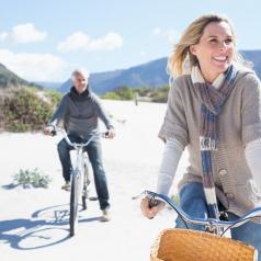 A middle-aged couple wearing warm clothes rides bikes along sandy beach, smiling and displaying enjoyment while looking off toward something in the distance