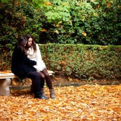 Two young people with long hair wearing coats and warm clothes sit on a bench in carpet of autumn leaves