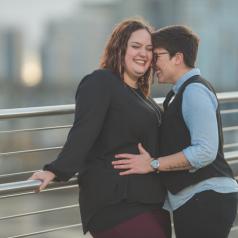 Young queer couple embrace happily while leanign on outdoor railing against bridge 