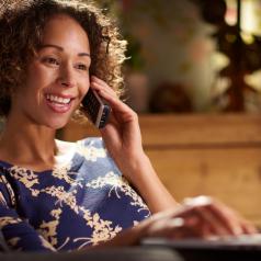 Person with short curly hair talks on cordless phone, smiling, while using laptop