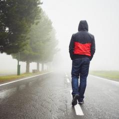 Rear view of person in black and red hooded sweatshirt walking alone down the middle of a road on a misty day