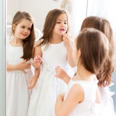 Two young girls with long hair wear white dresses and apply lip gloss in front of a mirror