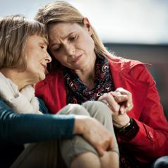 Mature woman and adult daughter sit close to each other with sad expressions on their faces. Younger woman is holding her mother