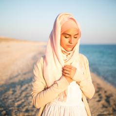 Woman on beach in cream-colored hijab clasps hands to chest in contemplative pose