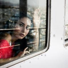 Adult with long dark hair wearing red top looks out train window with hand raised to wave goodbye