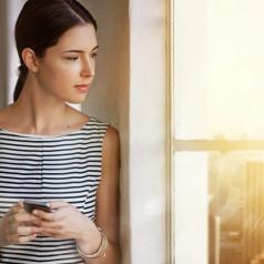 Person in business attire with hair tied back into ponytail holds phone and looks thoughtfully out window