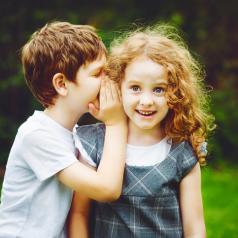 Child whispers into ear of smiling child with long curly hair
