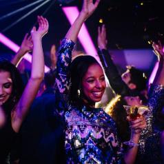 Happy young adult dancing with her friends while at a nightclub party with confetti, holding a drink on her hand