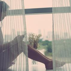 Young woman sitting on windowsill behind curtains and listening to the music