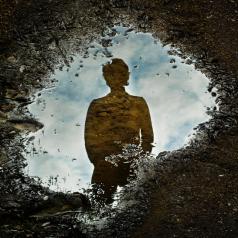 Puddle of water on ground reflects blue clouded sky and person with hands in pockets looking down into puddle
