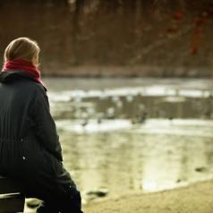 Person wearing red scarf and black coat sits on back of bench and looks out at lake