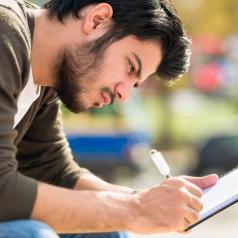 Close-up photo of young adult leaning over journal while sitting outdoors and writing 