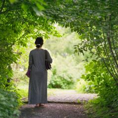 Adult in long gray wrap with hair back in clip walks through pathway under archway of green trees, standing alone, peacefully
