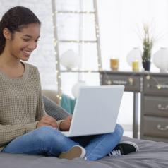 Teenager smiles while sitting on bed in decorated bedroom, legs crossed, using laptop