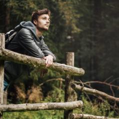 Young adult hiker with backpack in the forest leans on a wooden fence looking lost in thought