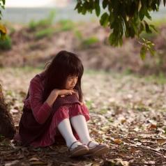 Distressed child with long dark hair sits under tree, knees drawn up to chest and hands on knees