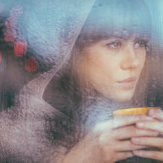 Person with long hair and bangs wearing hood holds mug close to face and looks out window on rainy day