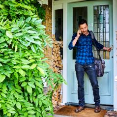 young adult in plaid shirt and jeans with leather bag speaks on phone while leaving house