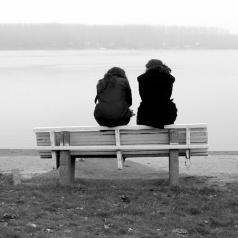 Photo shows rear view of two people with long hair wearing coats sitting on back of bench by water